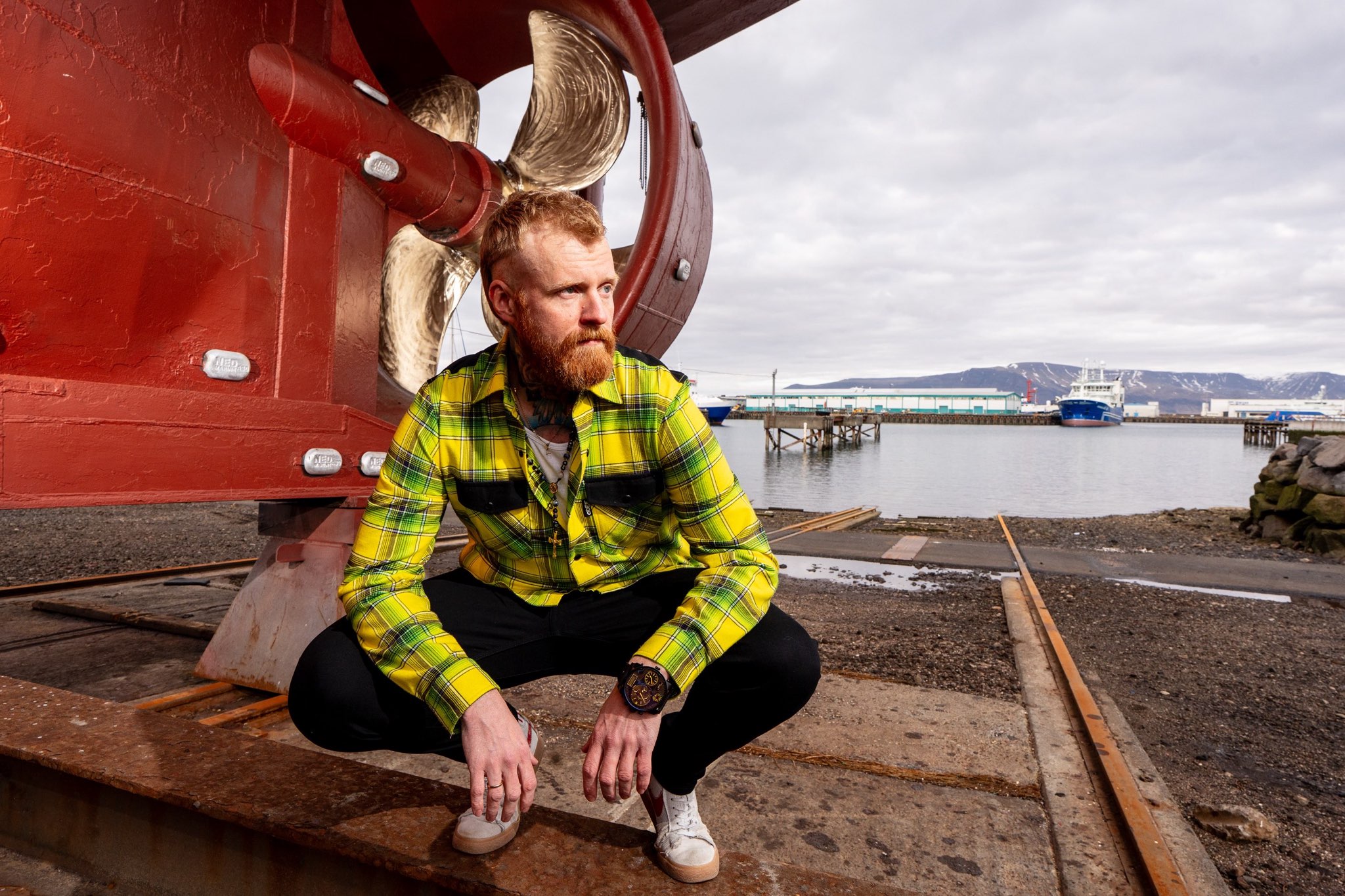 signal-2025-07-22-153608 a man with red hair wearing a bright yellow plaid shirt crouches down by a large red piece of machinery at a port. He looks off camera in an enigmatic way.